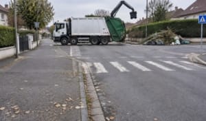 Un camion de collecte des déchets manœuvre à un croisement, avec des sacs de déchets en désordre sur le bord de la route dans un quartier résidentiel